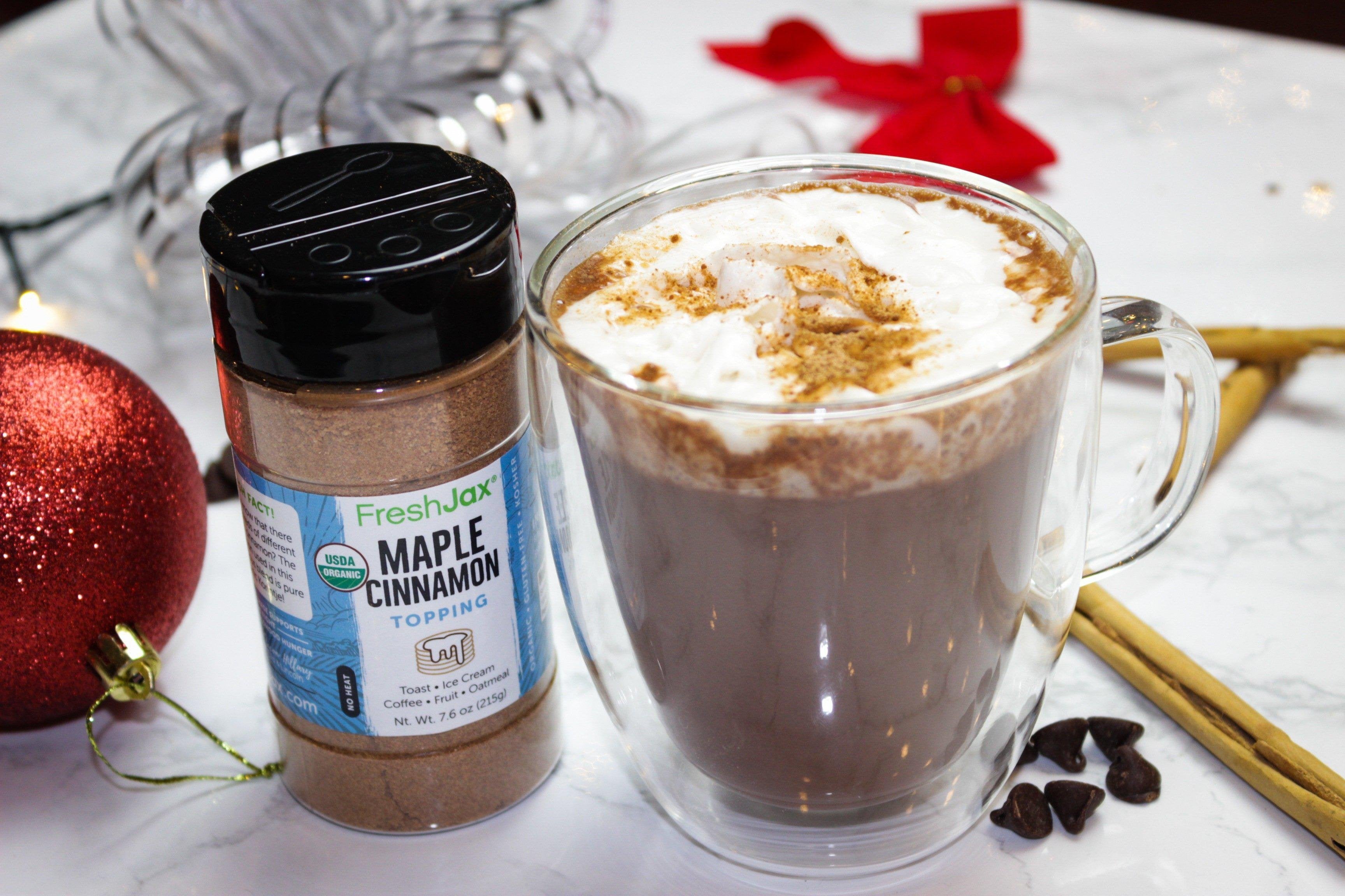 A large bottle of FreshJax Organic Maple Cinnamon Topping next to a mug of vegan hot cocoa topped with whipped cream and maple cinnamon. With a holiday themed background.