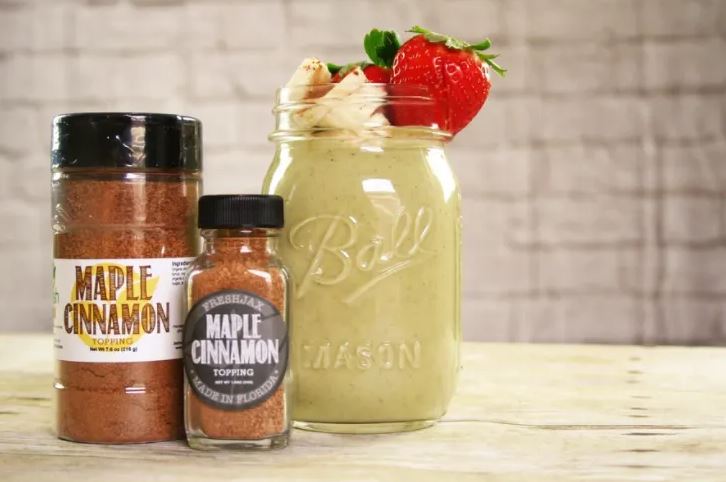 Two bottles of FreshJax Organic Maple Cinnamon Topping next to a Ball Jar filled with a maple cinnamon smoothie topped with whipped cream and strawberries.